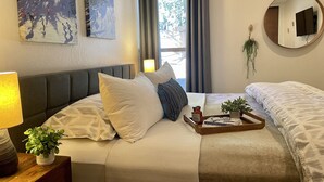 A hotel room with a bed, a bedside table with a lamp, a small plant, a tray with books and glasses, and a window with curtains.
