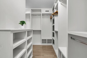 Un petit dressing étroit avec plusieurs étagères et une tringle à vêtements, une petite plante en pot et une grille d’aération au plafond.