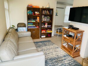 Ett vardagsrum med en vit lädersoffa, en träbokhylla fylld med brädspel, en tv och ett litet träbord med elektroniska apparater.