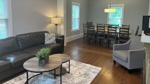 A living room with a dark leather sofa, a round coffee table, a gray armchair, a dining table with chairs, and a TV on a stand. - DOWNTOWN LAKE GENEVA 4 BR/4 BA House sleeps 15 (Lake Geneva)