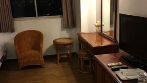A hotel room with a bed, a wicker chair, a small stool, a desk with a television, and a window with curtains.