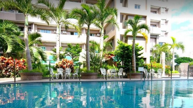 A swimming pool with clear water, surrounded by palm trees and a multi-story building with balconies.