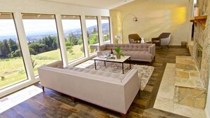 En rummelig stue med et stort vindue der giver udsigt over et grønt landskab, en beige sofa, et sofabord og en pejs.