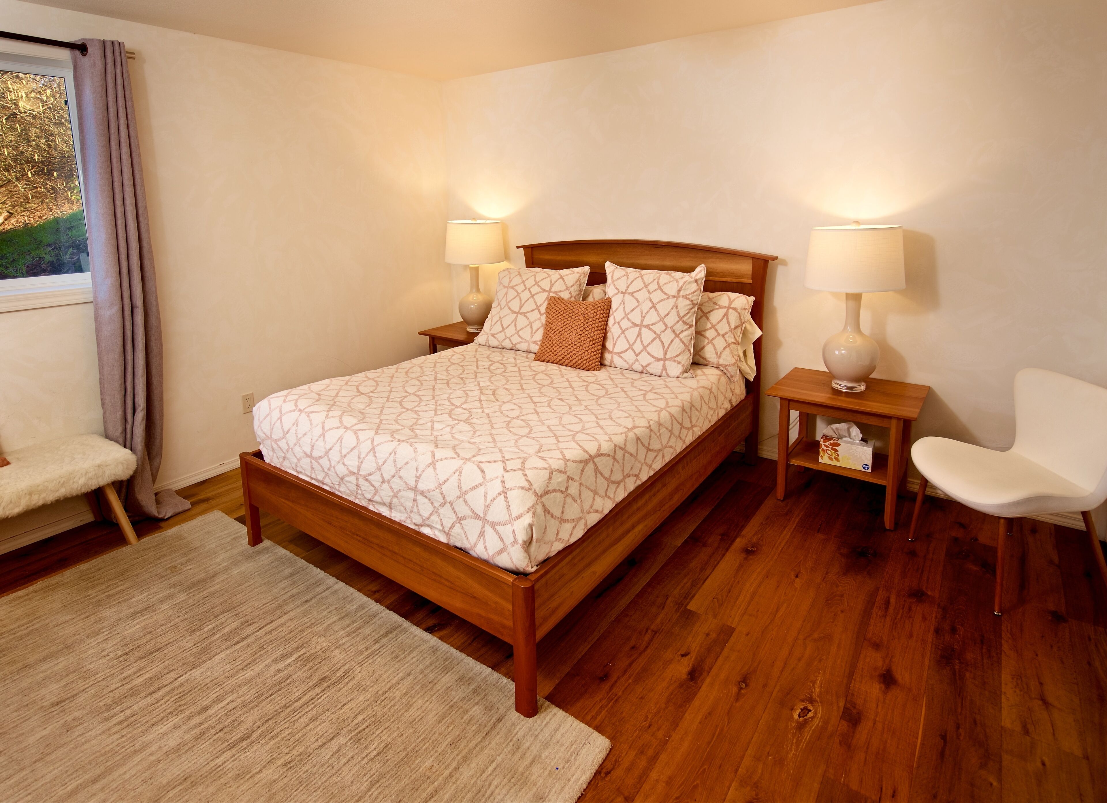 Un dormitorio con una cama de madera, dos mesitas de noche con lámparas, una silla blanca y una ventana con cortinas.