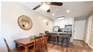 A dining area with a wooden table and chairs, a kitchen with white cabinets and appliances, and a ceiling fan.