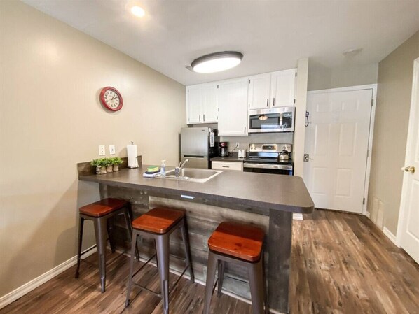 Una cocina con una isla central, taburetes de bar, armarios blancos, un microondas y un reloj en la pared.