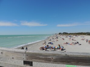 A beach with people sunbathing and swimming, a wooden fence in the foreground, and a clear blue sky.