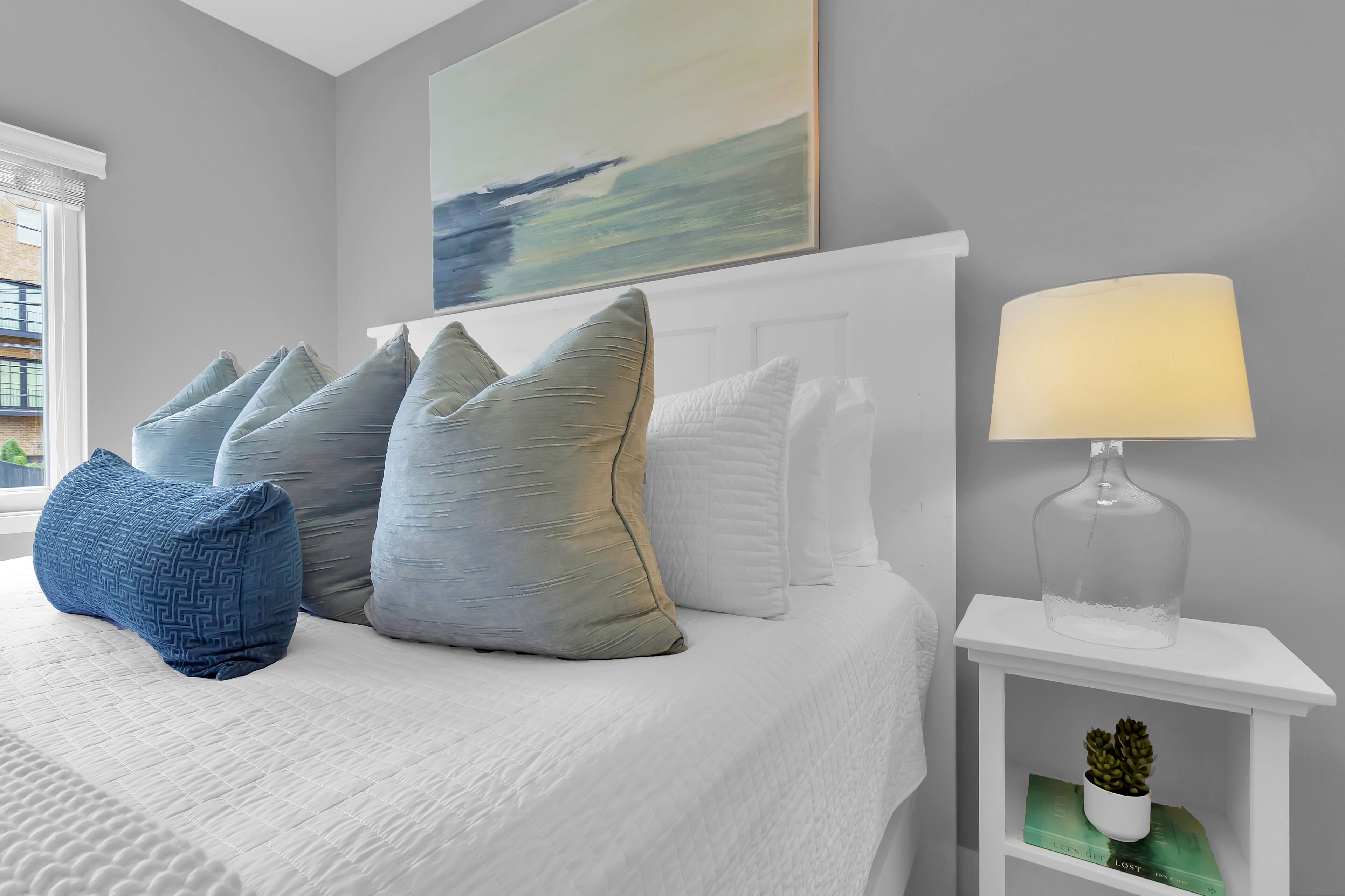 A neatly made bed with a white headboard, multiple pillows, a lamp on a side table, and a painting above the bed.