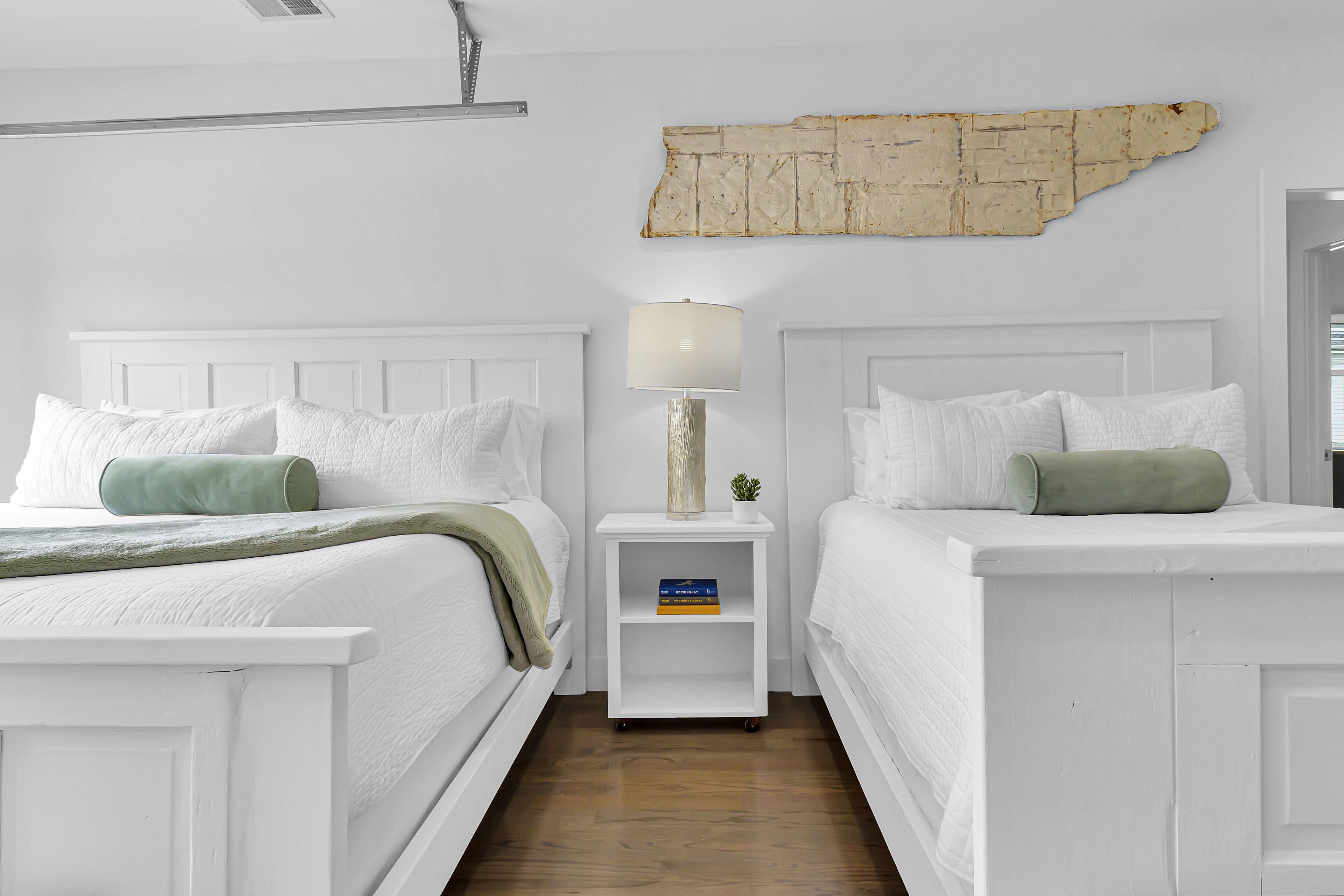 Une chambre d’hôtel avec deux lits, des têtes de lit blanches, une petite table de chevet avec une lampe, un élément décoratif mural et un coussin vert sur chaque lit.