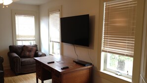 A living room with a wooden desk, a leather couch, a flat-screen TV mounted on the wall, and large windows with blinds. - Keweenaw Vacation Rental - Great for families and groups- sleeps up to 7 (Ahmeek)