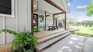 A covered porch with steps leading to a seating area, a small outdoor kitchen, and a view of a green landscape.