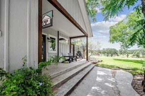 A covered porch with steps leading to a seating area, a sign reading "The Hideaway," and a view of a grassy landscape.