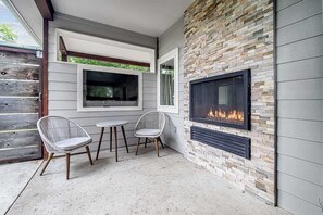 Un patio con dos sillas de mimbre, una mesita redonda y una chimenea de piedra con un televisor encima.