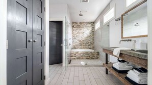Une salle de bain moderne avec un mur recouvert d’une mosaïque de pierre, un grand miroir et un banc en bois sur lequel sont posées des serviettes.