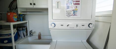 Une petite buanderie équipée d’un lave-linge et d’un sèche-linge blancs, d’un évier et d’une étagère contenant des produits de nettoyage.