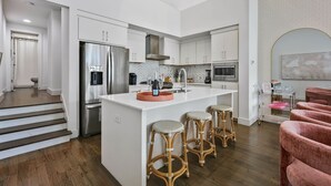A modern kitchen with white cabinetry, a stainless steel refrigerator, a central island with stools, and a dining area with pink chairs.