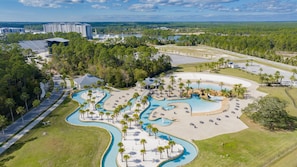 Vista aérea de um resort com várias piscinas, palmeiras e um edifício grande ao fundo.