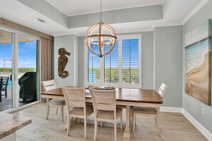 Uma sala de jantar com mesa e cadeiras de madeira, um candelabro e vista para o exterior através de grandes janelas.