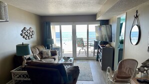 Een woonkamer met een bank, salontafel en televisie, die uitkomt op een balkon met uitzicht op zee.