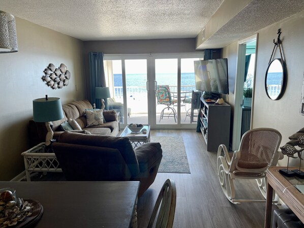 A living room with a sofa, coffee table, and TV, leading to a balcony with a view of the sea.