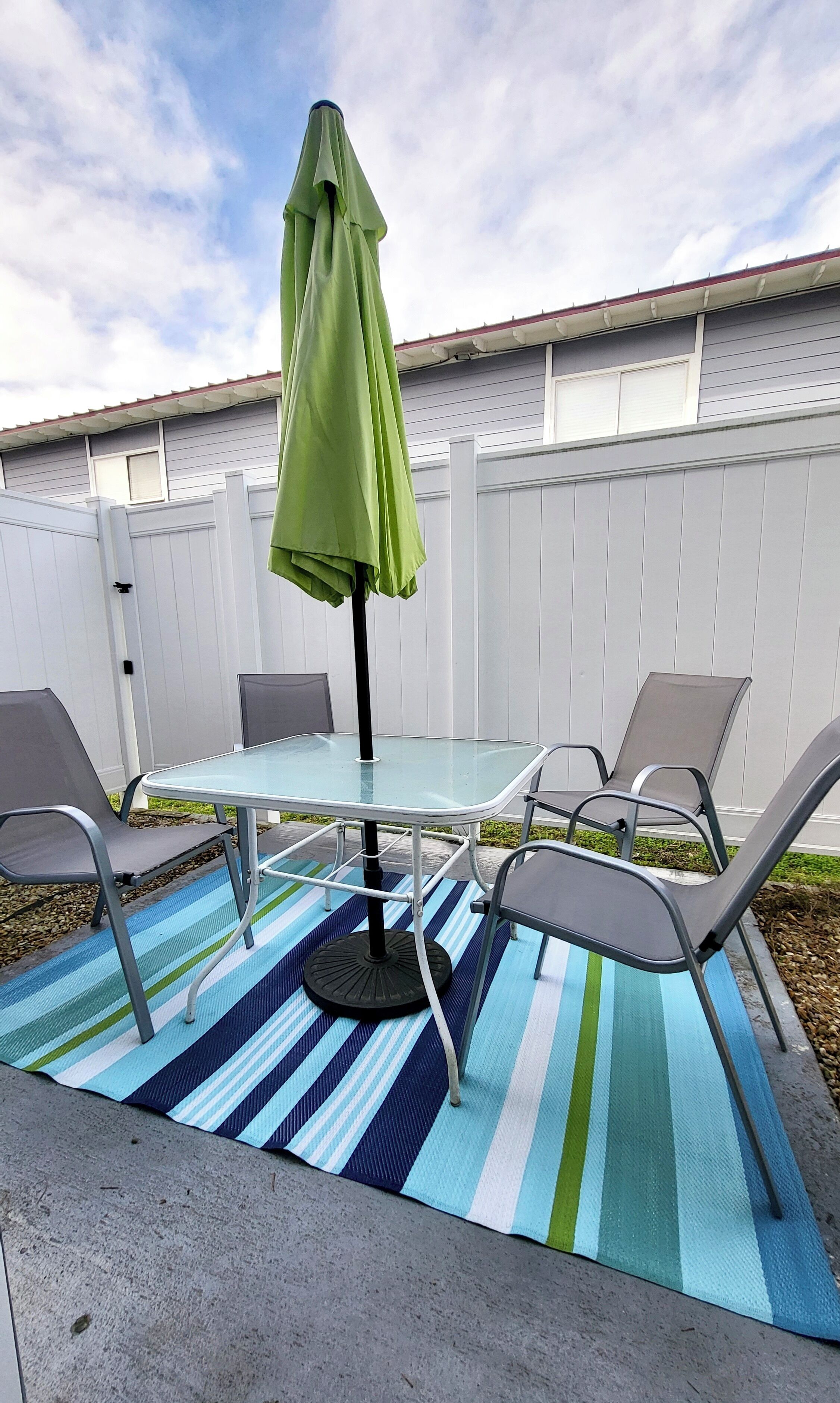 Une terrasse extérieure avec une table en verre et des chaises en métal, un parasol vert et un tapis à rayures.
