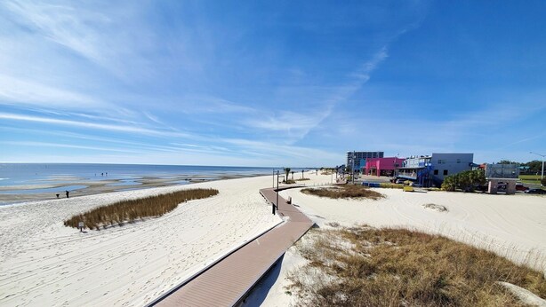 Een strand met een houten looppad, een zandpad en gebouwen op de achtergrond.