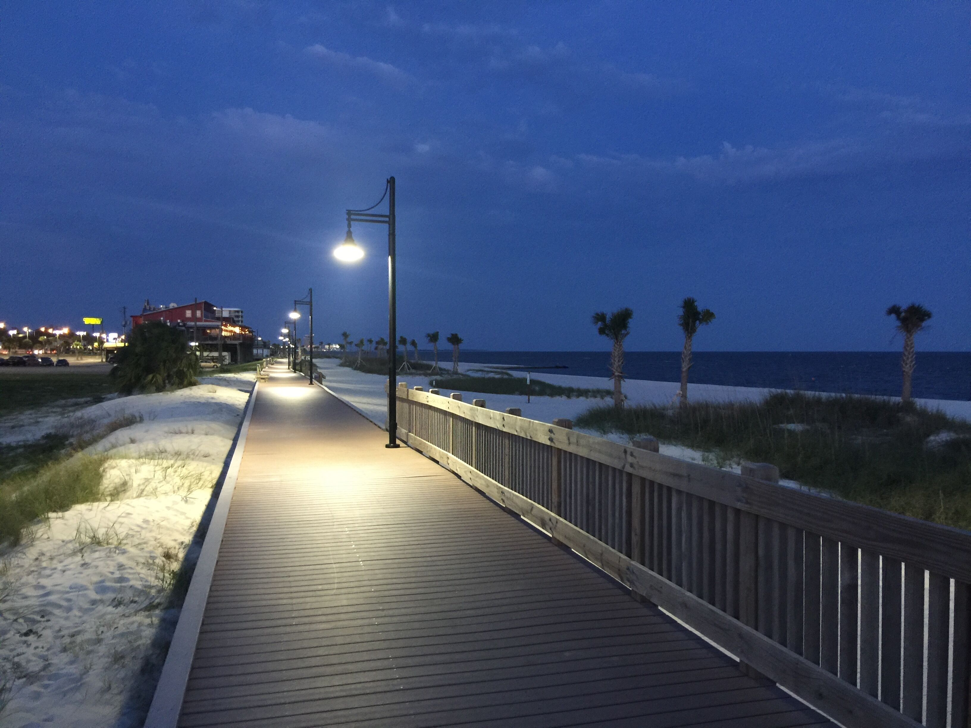 A wooden boardwalk with street lamps, leading to a beach with palm trees and the ocean.