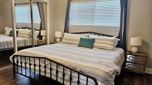 Een slaapkamer met een groot bed, een spiegel, een nachtkastje en een raam met jaloezieën.