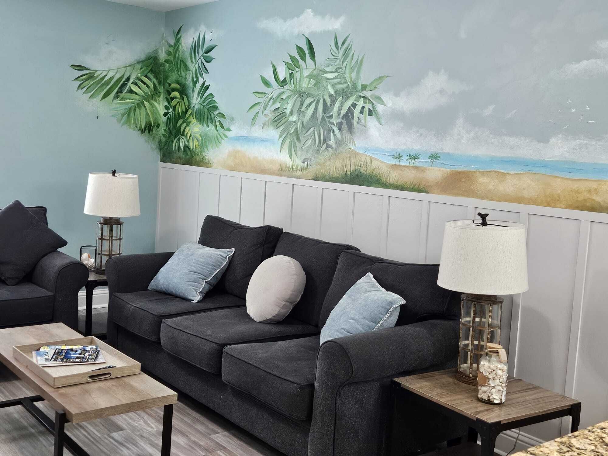 Een woonkamer met een donkergrijze bank, twee bijzettafels met lampen, een salontafel en een muurschildering van een strand.