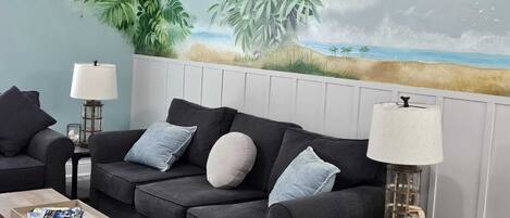 Een woonkamer met een donkergrijze bank, twee bijzettafels met lampen, een salontafel en een muurschildering van een strand.