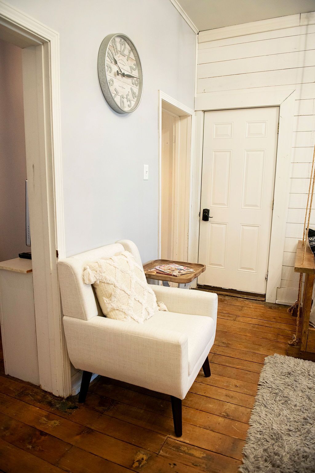 Un salon avec un fauteuil blanc, un plancher en bois, une porte blanche et une horloge murale.