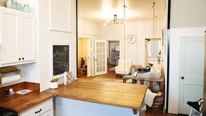 A kitchen with white cabinets, a chalkboard with 'Happy Spring', a wooden counter, and a view into a dining area with a white chair and a hanging light fixture. - The Farmhouse Loft "Greene County's Guesthouse" (Carrollton)