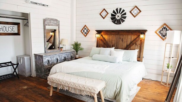 A bedroom with a wooden headboard, a large bed with white bedding, a bench, and a dresser with a mirror. - The Farmhouse Loft "Greene County's Guesthouse" (Carrollton)
