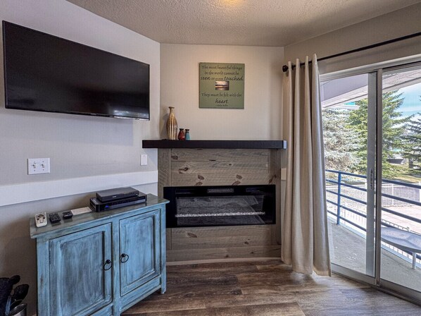 A room with a flat-screen TV mounted on the wall, a wooden cabinet with a television, a fireplace, and a large sliding glass door leading to an outdoor area.