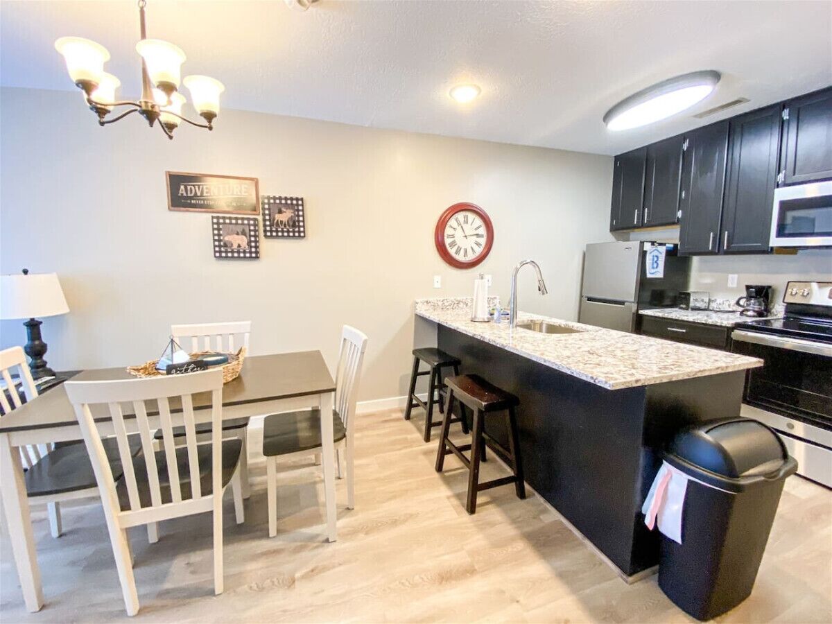 A modern kitchen with a central island, white chairs, a dining table, a clock, and a microwave.