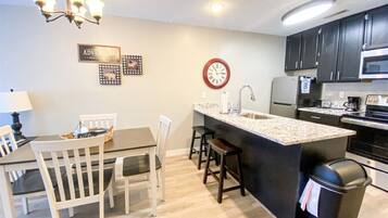 A modern kitchen with a central island, white chairs, a dining table, a clock, and a microwave.