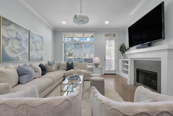 A modern living room with a sofa, ottomans, a coffee table, a flat-screen TV, and a fireplace.