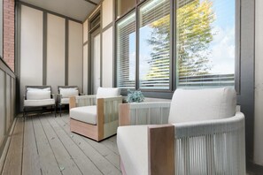 A sunlit balcony with wicker furniture, large windows, and a view of trees.
