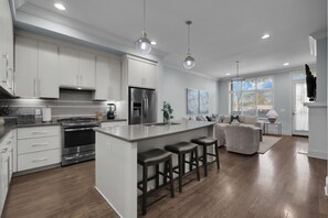 A modern kitchen with stainless steel appliances, a central island with bar stools, and a dining area with a sofa and a TV.