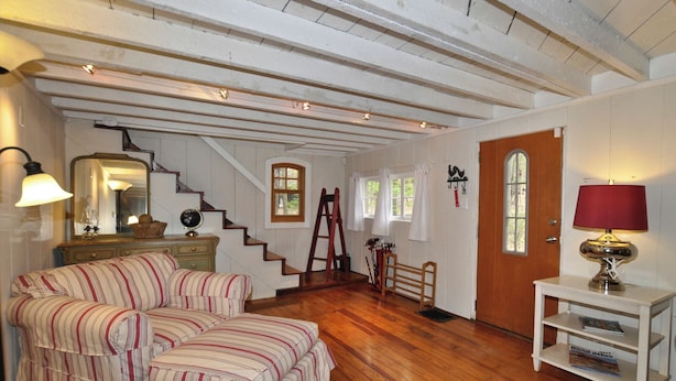 Une pièce chaleureuse avec un plancher en bois, un canapé à rayures, un escalier, une porte en bois et une petite table avec une lampe.