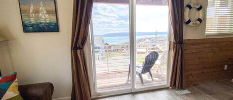 Una habitación con un gran ventanal corredizo que da a una terraza con vista al mar. Hay un cuadro en la pared, una silla y una alfombra en el suelo.