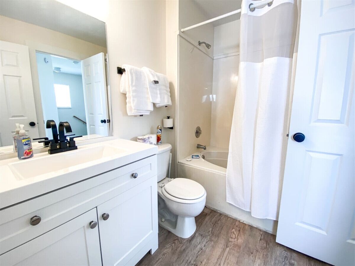 A bathroom with a white sink, toilet, and shower. There are towels hanging on the wall and a bottle of hand soap on the sink.