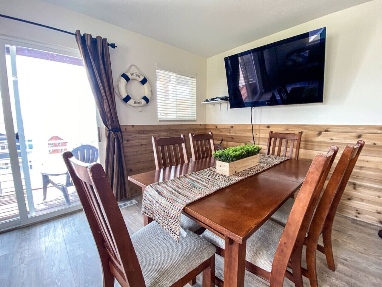 A dining area with a wooden table, six chairs, a television mounted on the wall, and a view of the outside through a sliding door.