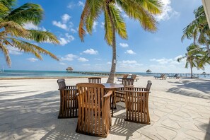 Een eetgedeelte aan het strand met houten tafels en stoelen, palmbomen en uitzicht op de oceaan en de pier.