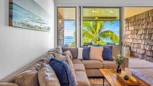 A modern living room with a large sectional sofa, a wooden coffee table, and a view of palm trees through large windows. - Beachfront 2BD Unobstructed Ocean Views - Kulakane 304 (Lahaina)