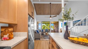 A modern kitchen with wooden cabinets, a large island, and a view of the living area with a sofa and a plant. - Beachfront 2BD Unobstructed Ocean Views - Kulakane 304 (Lahaina)