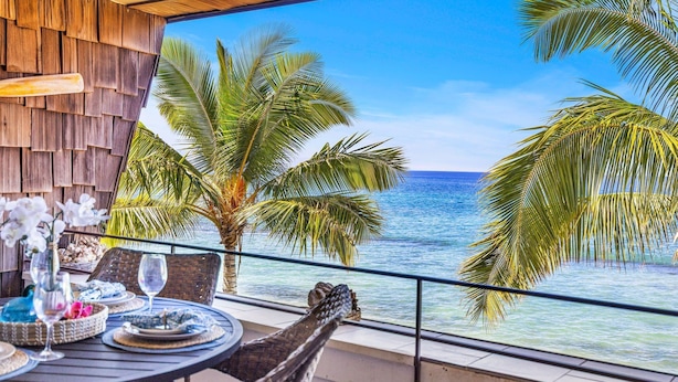 A balcony with a view of the ocean, palm trees, and a dining table set with plates and glasses. - Beachfront 2BD Unobstructed Ocean Views - Kulakane 304 (Lahaina)