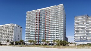 A high-rise hotel with multiple balconies, a central red structural feature, and a clear blue sky. - Last Minute Rate, Direct Oceanfront 4BR/3BA, Sleep 16, Central Location (Myrtle Beach)