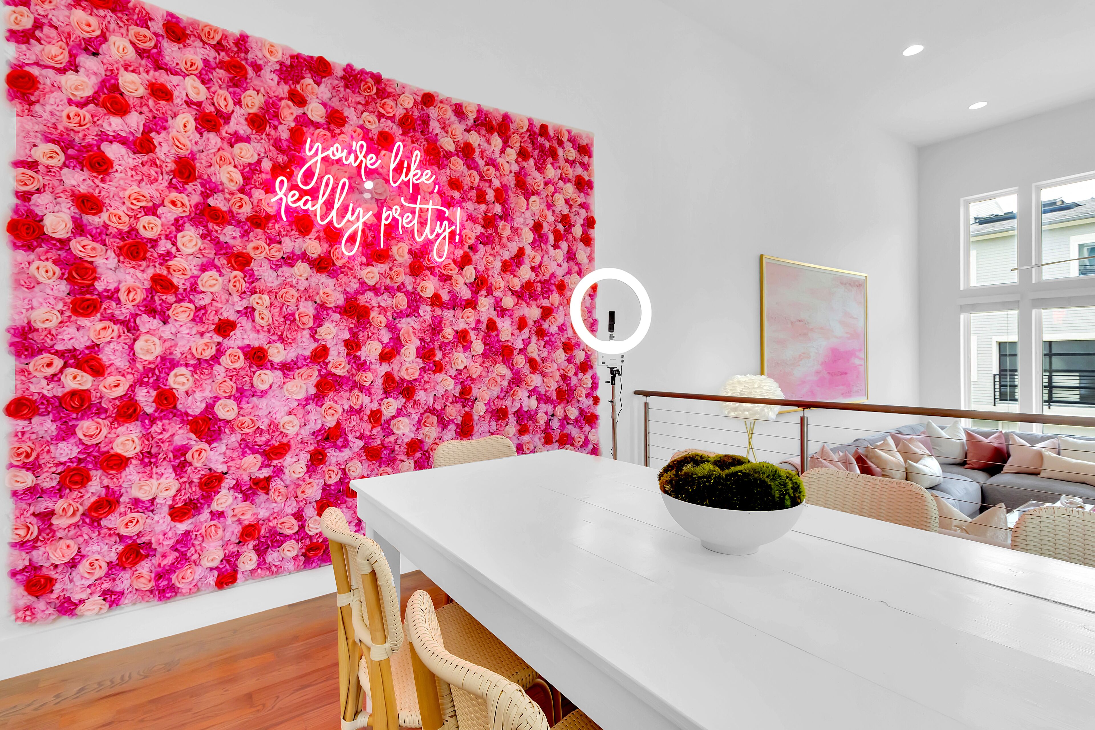 A modern living room with a white dining table, wicker chairs, a sofa with cushions, a large floral wall, and a ceiling light fixture.