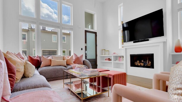 A modern living room with a sectional sofa, a glass coffee table, a flat-screen TV mounted above a fireplace, and large windows offering a view of the neighborhood.